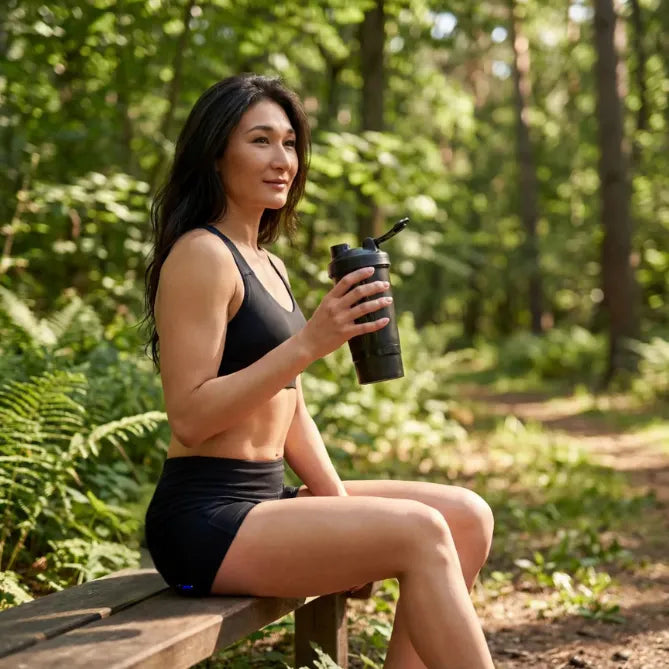 Woman sitting on a wooden bench in a forest, holding a black shaker bottle.